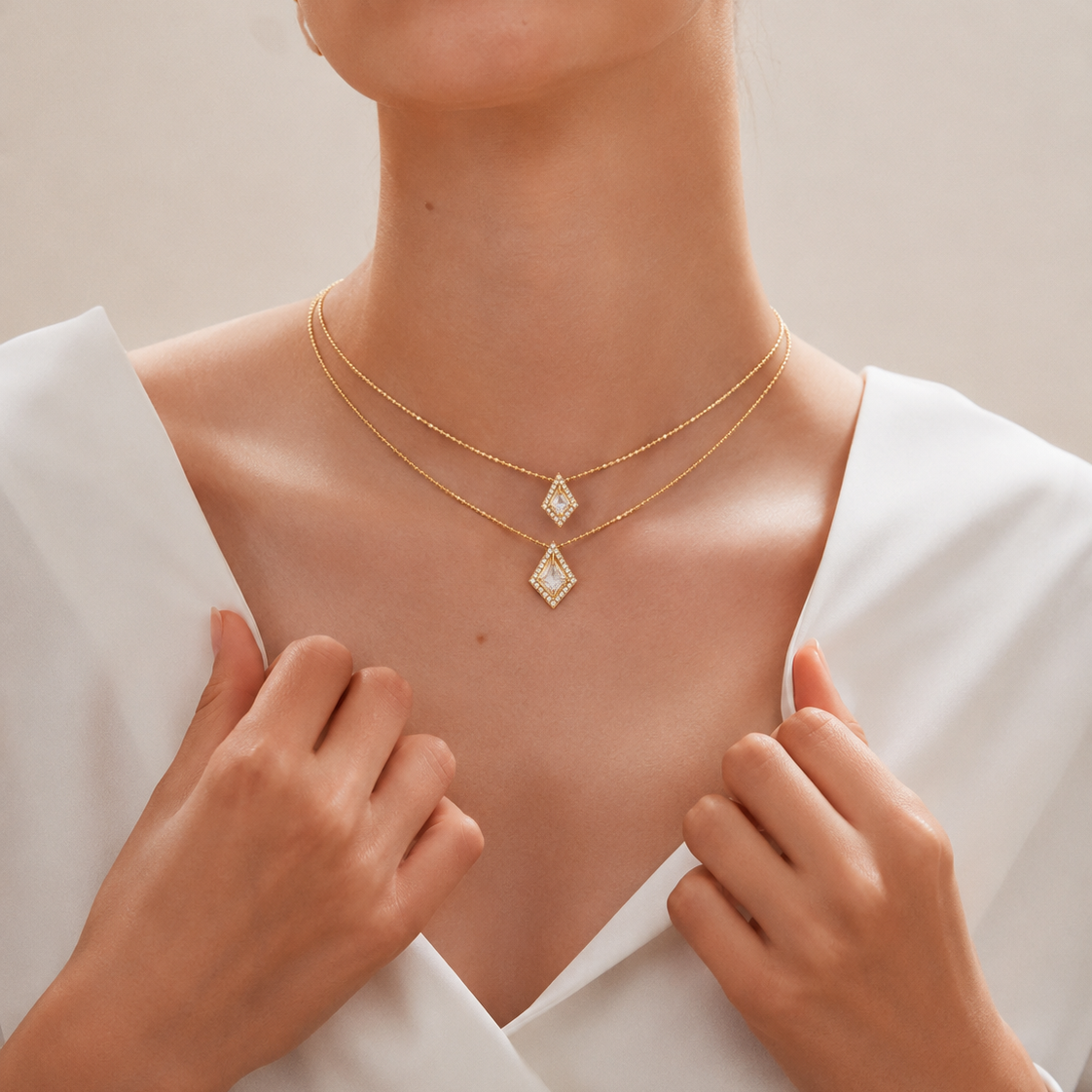 Two layered yellow gold ball-chain necklaces resting on white silk, each featuring a diamond-shaped rhombus pendant with a central square gemstone surrounded by smaller diamonds.